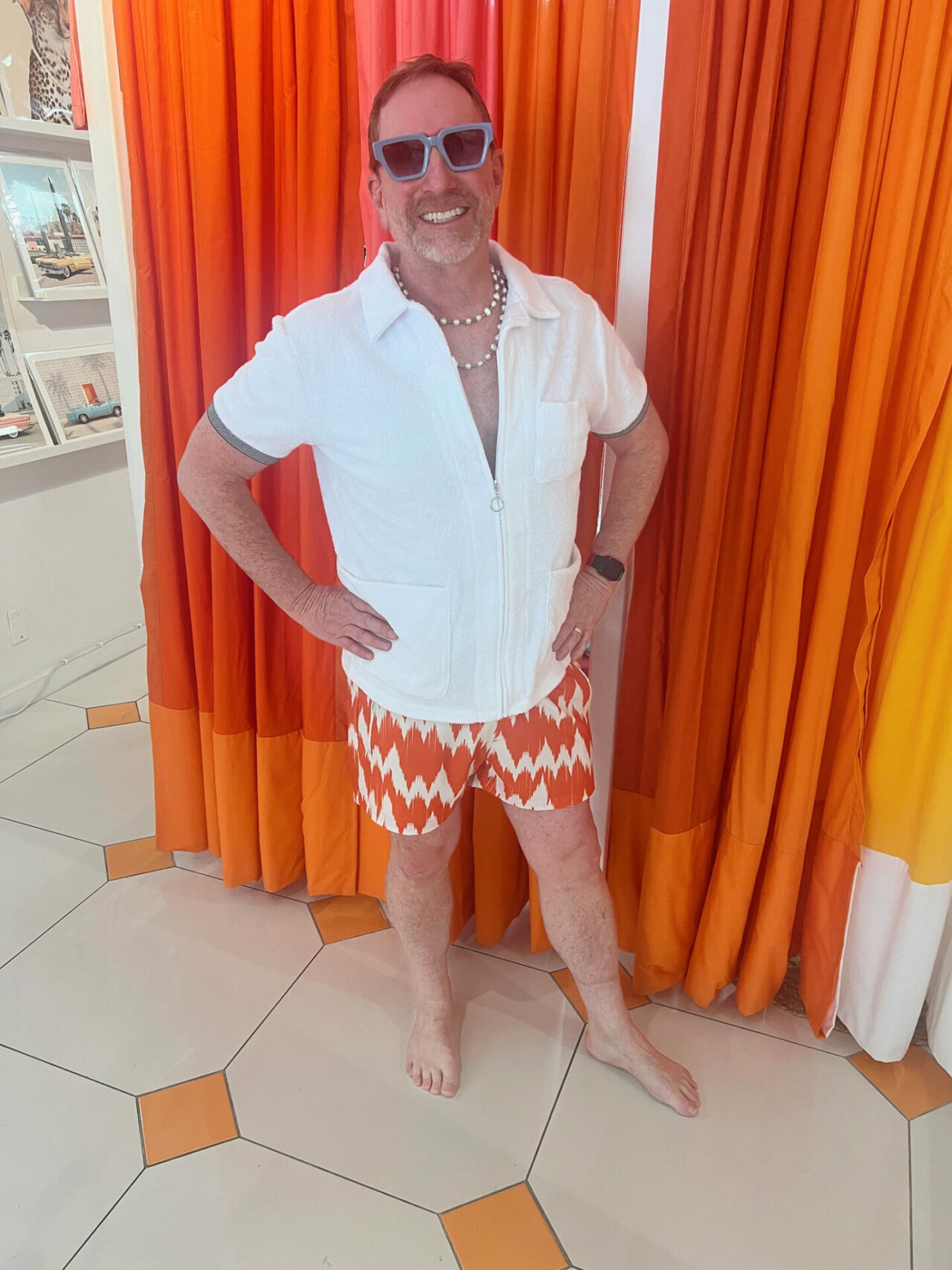 Eric wearing an outfit from El Tuggle in Palm Springs behind brigh orange changin curtains and a pearl necklace